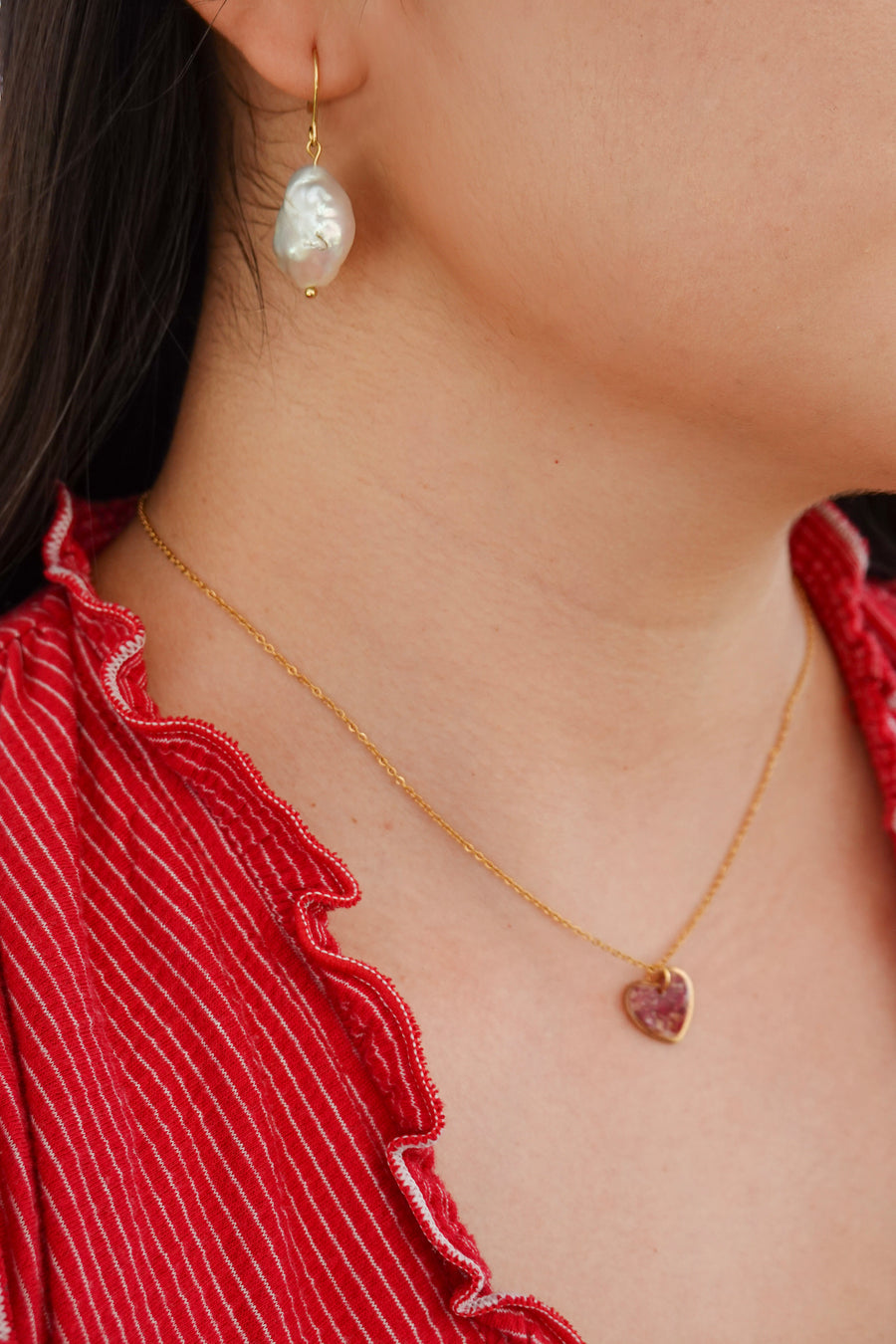 Close-up of a person wearing a gold necklace with a heart-shaped crushed rose petals pendant and pearl earrings.